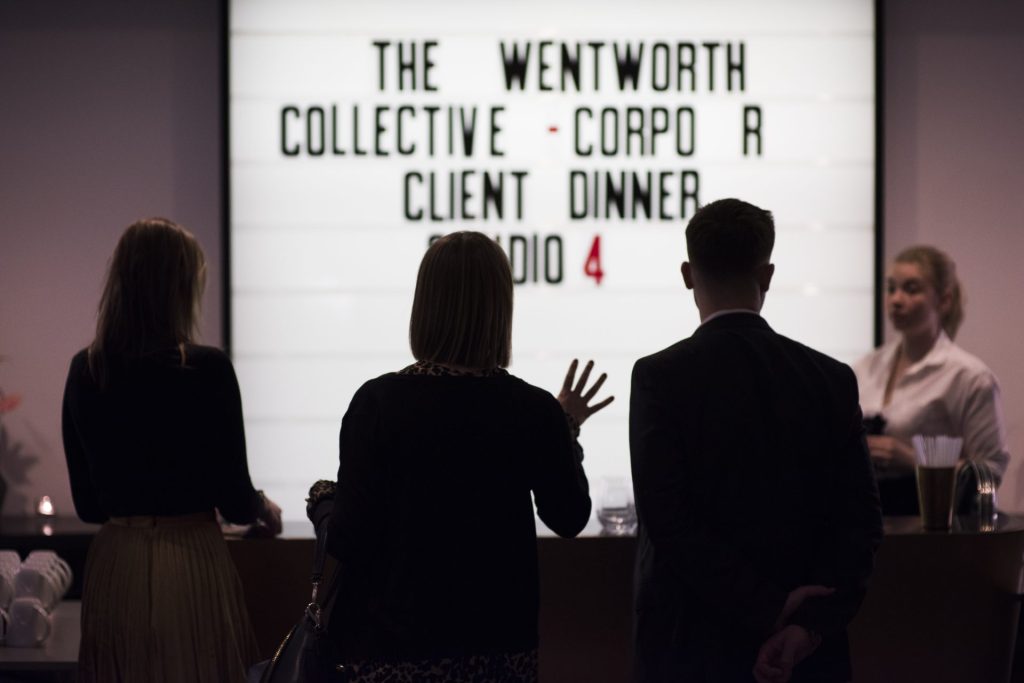 We can see the back of The Wentworth Collective founder, Lisa, during event planning, discussing plans with team, in front of a cinema-style light box announcing the event.
